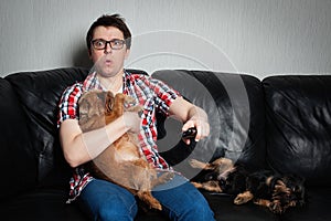 Closeup portrait, young man in the red shirt, sitting on black leather couch with two dogs, watching TV, holding remote, surprised