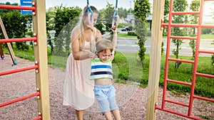 Closeup portrait of young mother helping and holding her little son hanging and swinging on sportrs rings at palyground