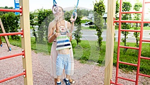 Closeup portrait of young mother helping and holding her little son hanging and swinging on sportrs rings at palyground