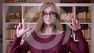 Closeup portrait of young redhead attractive caucasian female student in glasses having her fingers crossed with anxiety