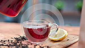 Closeup pouring fresh hot aroma black tea from jar teapot into glass cup on wooden table slowmo