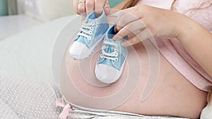 Closeup of pregnant woman holding shoes for newbon babies on her big belly. Concept of pregnancy, preparing and