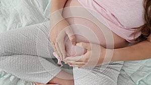 CLoseup of pregnant woman sitting on bed and making shape of heart with hands on her big belly. Concept of expecting