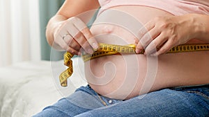 Closeup of pregnant woman sitting on bed and measuring her big belly with measuring tape