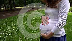 Closeup of pregnant woman stroking her big belly standing in park meadow