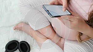 Closeup of pregnant womansitting on bed and typing message on smartphone after listening music in headphones. Concept of