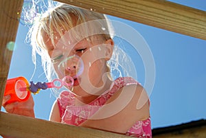 Closeup of a pretty, young girl wearing a pink dress, blowing bubbles from the top of her climbing frame, on a sunny summers day