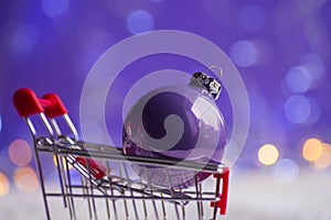 Closeup purple Christmas ball in small supermarket trolley with garland lights on lilac bokeh background.