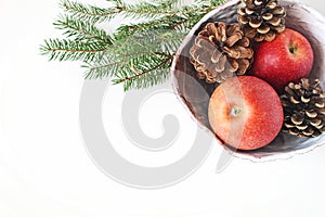 Closeup of red apple fruit and pine cones in ceramic bowl and spruce tree branches on white table background. Christmas