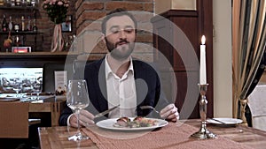 Closeup of restaurant guest. Well dressed, confident young man with a beard is eating plate with meat dish in fine dining restaura