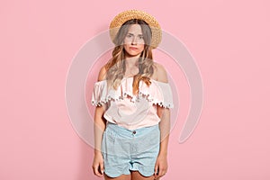 Closeup of sad pensive young woman with long wavy hair and straw hat, wearing summer clothes, looks unhappy, feels bad, looking