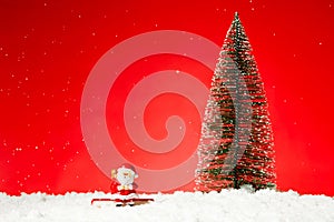 Closeup of a Santa Claus toy on fae snow with a Christmas tree on it against a red background