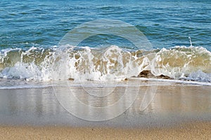 Closeup of sea wave with white foam comes ashore. The sea wave is reflected in the yellow wet coastal sand