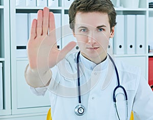 Closeup on serious male medical doctor showing stop gesture at office background.