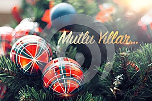 Closeup of shiny red bauble hanging from a decorated Christmas tree. Mutlu Yillar means Happy new year