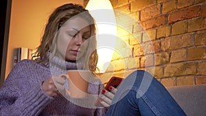 Closeup shoot of adult caucasian blonde female browsing on the phone while sitting on the couch laidback and drinking