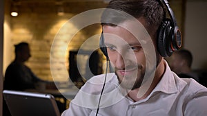 Closeup shoot of adult caucasian businessman in headphones texting on the tablet and smiling cheerfully on the workplace