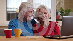 Closeup shoot of senior happy couple using the laptop on the desk with cups with tea indoors in a cozy apartment in a