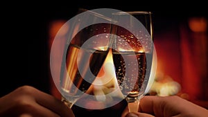Closeup shoot of two hands of friends clinking glasses full of champagne chilling indoors with cozy warm fireplace on