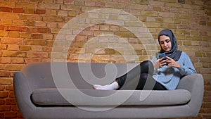 Closeup shoot of young attractive muslim female in hijab messaging on the phone while resting laidback on the couch