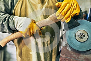 Closeup shot of an adult craftsman manufacturing in his workshop. Professional senior worker man grinding the hatchet with sparks