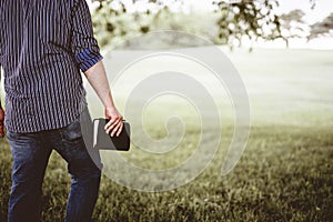 Closeup shot from behind of a male walking on a grassy field while holding the Bible