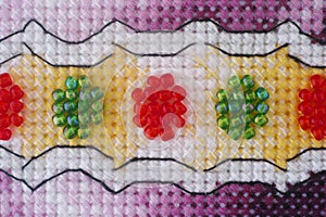 A closeup shot of a cross stitch mades with purple, yellow and white threads and red and green beads