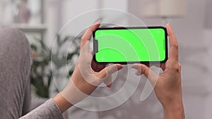 Closeup shot of a girl lying on a sofa using a smartphone in a horizontal position with a green screen mockup in a