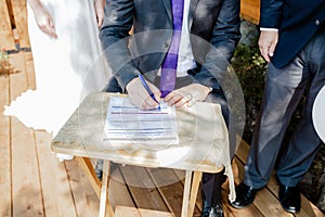 Closeup shot of a groom signing a wedding contract papers on a wooden table on a sunny day