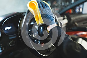 Closeup shot of a hand in black glove holding orange microfibre cloth and wiping the steering wheel of a car during car