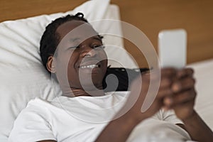 Closeup Shot Of Happy Young Black Man Using Smartphone While Lying In Bed