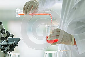 Closeup shot of orange red reagent in glass volumetric cylinder poured in beaker by scientist in white lab coat and rubber gloves