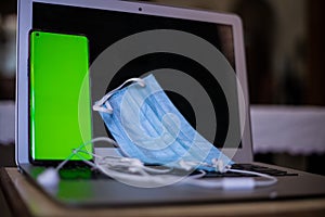 Closeup shot of a phone with a greenscreen, earphones, and a face mask on a laptop