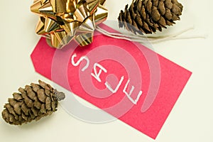 Closeup shot of a red sale tag with two pine cones and a gold pull bow on white background