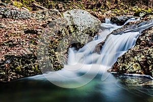 Closeup shot of a small water cascade in a stream surrounded by stones and autumnal leaves