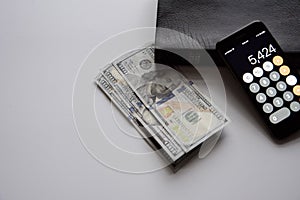 Closeup shot of a smartphone and hundred dollar bills near a book on a white surface