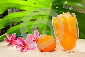 Closeup shot of a transparent glass full of orange juice next to pink plumerias