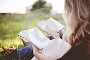 Closeup shot of two people sitting in a grassy field and reding the bible with blurred background