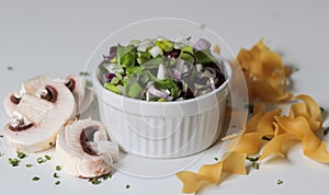 Closeup shot of a white bowl of a vegetable salad, sliced mushrooms and raw pasta on a white surface