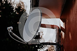 Closeup shot of a white satellite dish on fixed on a red wall with trees on the background