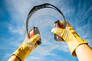 Closeup shot of yellow gloved hands holding red noise canceling construction headphones to blue sky