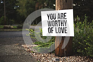 Closeup shot of You are worthy of love signage in front of a tree log near the road photo