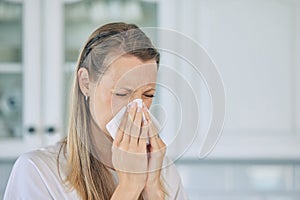 Closeup of sick caucasian woman holding tissue and blowing her nose while at home. Woman suffering from seasonal allergy