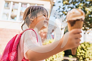 Closeup side view of cute little girl smiling and walking along city street and eating ice cream outdoor. Happy kid girl looks to