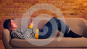 Closeup side view portrait of young pretty girl using the phone and laughing cheerfully lying on the couch laidback in a