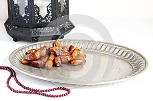 Closeup of silver plate with date fruits, prayer beads and ornamental dark Moroccan, Arabic lantern on the white table