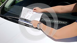 Closeup slow motion video of young woman putting for sale sign under the screen wipers of her car. Woman selling her