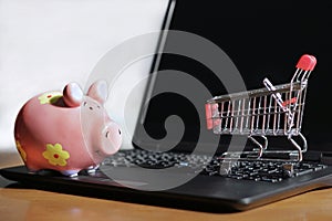 Closeup of a small shopping cart and a piggy bank on a laptop on the table - online shopping concept