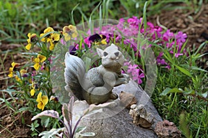 Closeup of a small squirrel statue on a rock in a garden covered in flowers