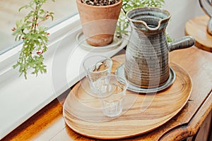 Closeup of small water cups next to a coffee pot, jezve in a wooden serving tray next to house plant